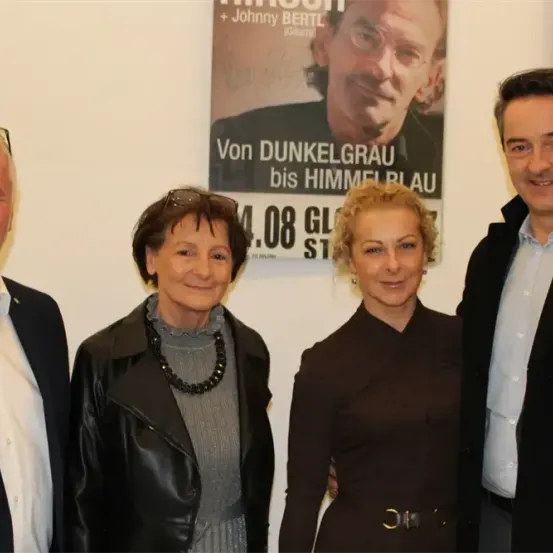 Vier Personen, zwei Männer und zwei Frauen, posieren für ein Foto vor einem Poster mit einem lächelnden Mann und dem Text 'Von Dunkelgrau bis Himmelblau'.