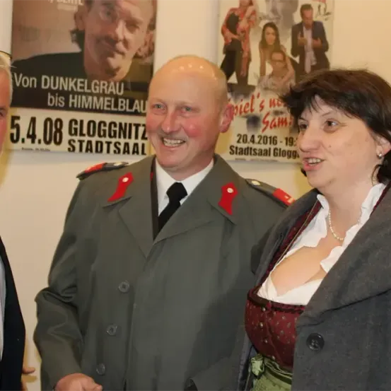 Drei Personen, zwei Männer und eine Frau, stehen vor einem Plakat, das eine Veranstaltung ankündigt.