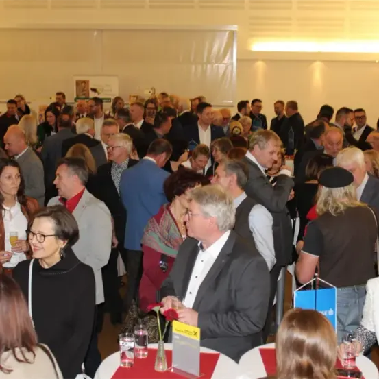 Eine Gruppe von Menschen, sowohl Männer als auch Frauen, formell gekleidet, versammelt sich in einem Saal. Sie stehen und reden, einige halten ein Glas mit Getränken. Ein Tisch in der Mitte hat eine Blumenvase, ein gelbes Schild und eine blaue Tasche.