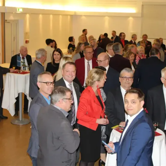 Eine Gruppe von Menschen in formeller Kleidung versammelt sich in einem Raum. Einige halten Getränke in den Händen. Sie scheinen zu sprechen und lächeln. Es gibt Tische mit weißen Tischdecken.