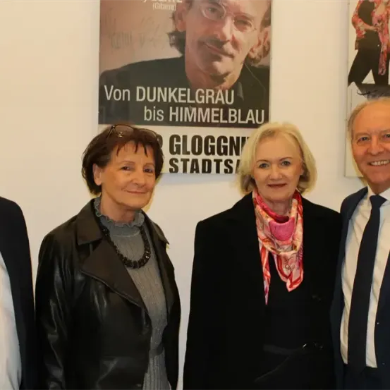 Vier Menschen stehen vor einer Wand mit einem Plakat. Der Mann auf der rechten Seite trägt einen Anzug. Die beiden Frauen tragen Schals. Die Frau auf der linken Seite trägt eine Halskette. Der Mann auf der linken Seite trägt eine Brille.