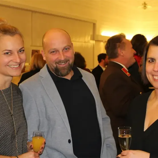 Drei Menschen stehen zusammen, lächeln und halten Getränke in der Hand. Die Frau auf der linken Seite hält ein Glas Orangensaft, während die anderen beiden Champagnergläser haben. Hinter ihnen stehen andere Menschen, möglicherweise auf einer Party.