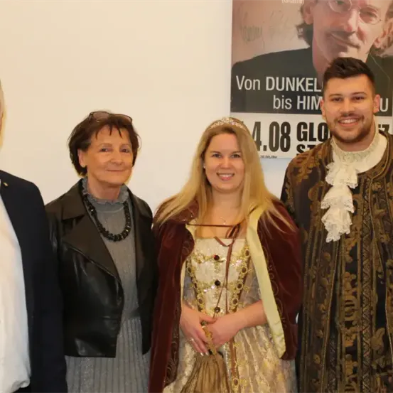 Vier Personen stehen vor einem Plakat für eine Theateraufführung. Die Frau in einem goldenen Kleid hält eine Handtasche.