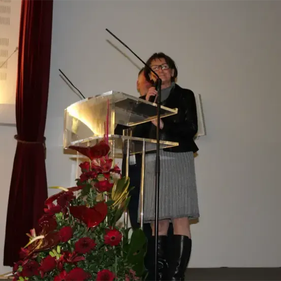 Eine Frau mit Brille steht hinter einem Rednerpult mit Mikrofonen und hält ein Mikrofon in der Hand. Sie lächelt und scheint eine Rede zu halten. Das Podium ist mit roten Blumen geschmückt. Hinter ihr steht eine Person. Die Wand ist weiß und eine rote Gardine befindet sich links.