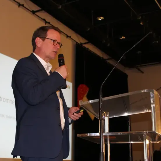 Ein Mann in Anzug und Brille steht an einem Podium, hält ein Mikrofon in der Hand und spricht hinein, mit einer Projektionsleinwand im Hintergrund.