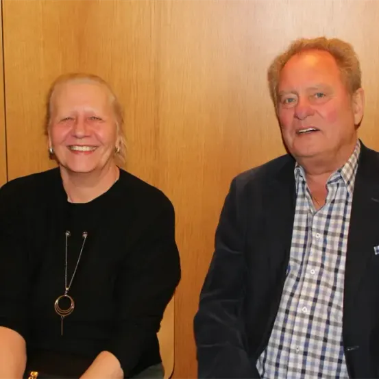 Eine ältere Frau und ein Mann sitzen nebeneinander und lächeln für die Kamera. Die Frau trägt ein schwarzes Shirt und eine Halskette. Der Mann trägt ein kariertes Hemd unter einem schwarzen Jackett. Sie stehen vor einer Holzwand.