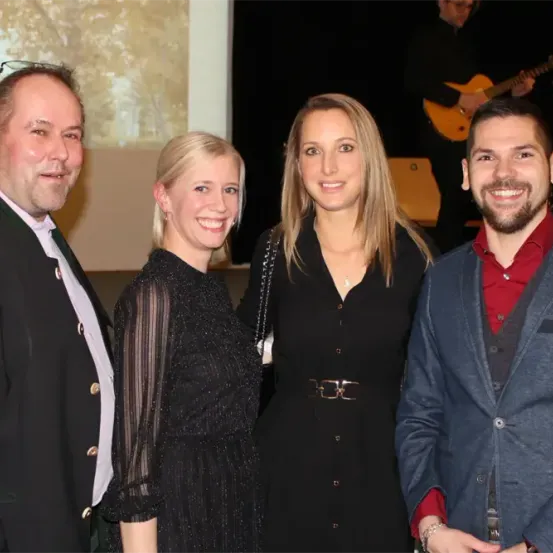 Vier Menschen posieren für ein Foto vor einer Kulisse mit einem großen Baum. Der Mann links trägt einen schwarzen Anzug. Die beiden Frauen in schwarzen Kleidern lächeln, eine hält eine Handtasche. Der Mann rechts trägt ein rotes Hemd und einen blauen Blazer. Im Hintergrund spielt ein Mann Gitarre.