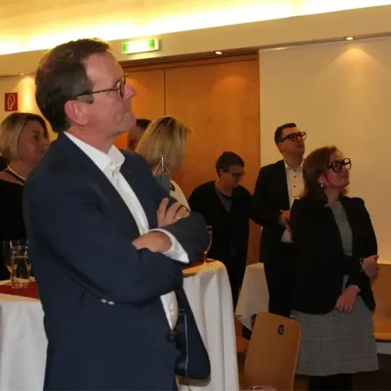 Eine Gruppe formell gekleideter Menschen steht in einem Raum, wahrscheinlich bei einer Veranstaltung. Ein Mann mit Brille schaut nach oben, während andere in die Ferne schauen. Einige Tische mit Gläsern und Stühlen sind in der Nähe. Eine Lampe ist an der Decke befestigt.