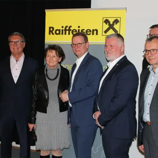 Eine Gruppe von sechs Erwachsenen, drei Männer und drei Frauen, gekleidet in formeller Kleidung, posiert für ein Foto vor einem Raiffeisen-Banner.