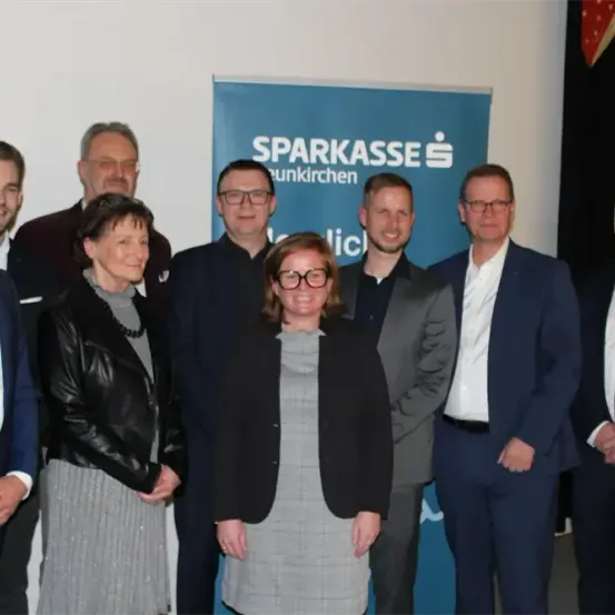 Eine Gruppe von Menschen in formeller Kleidung steht vor einem blauen Banner mit dem Text 'Sparkasse.' Sie lächeln und scheinen für ein Foto zu posieren.
