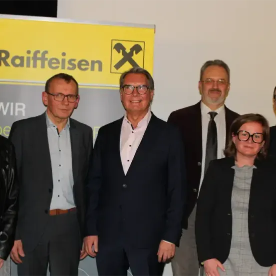 Eine Gruppe von Personen in formeller Kleidung steht vor einem Raiffeisen-Banner. Das Banner zeigt das Raiffeisen-Logo und den Schriftzug 'Raiffeisen' in großen schwarzen Buchstaben.