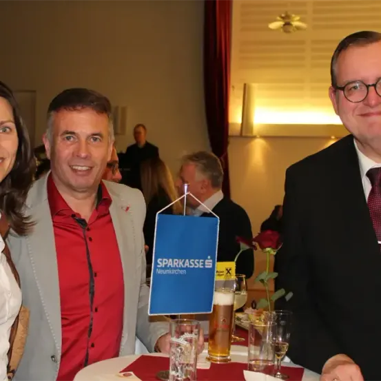 Drei Menschen lächeln vor einem Tisch mit Getränken. Ein Mann in Anzug steht neben einer Frau und einem anderen Mann. Ein Banner mit dem Text 'SPARKASSE' hängt hinter ihnen.
