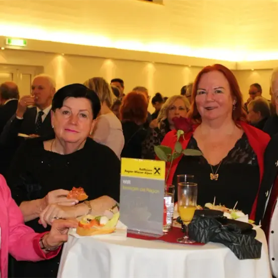 Eine Gruppe von Erwachsenen versammelt sich in einem Esszimmer. Drei Frauen sind sitzend und ein Mann steht hinter ihnen. Es gibt einen Tisch mit Essen und Getränken. Ein Schild auf dem Tisch lautet 'Wir Verweilen die Region'.