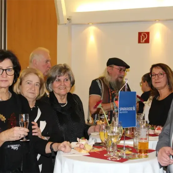 Eine Gruppe älterer Menschen sitzt bei einer Veranstaltung um einen Tisch herum, lächelt und hält Gläser mit Wein und Bier in den Händen. Der Tisch ist mit einer weißen Tischdecke bedeckt und hat verschiedene Getränke und Speisen darauf. Im Hintergrund befindet sich ein Schild mit dem Wort 'Sparkasse'.
