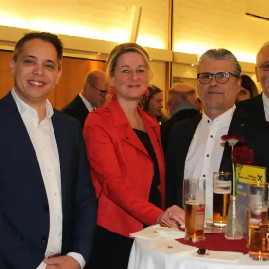 Vier Erwachsene, zwei Männer und zwei Frauen, stehen eng beieinander, lächeln und posieren für ein Foto auf einer Party. Der Mann links trägt einen dunklen Anzug, und die Frau rechts trägt eine rote Blazerjacke. Vor ihnen steht ein Tisch mit Gläsern Bier.