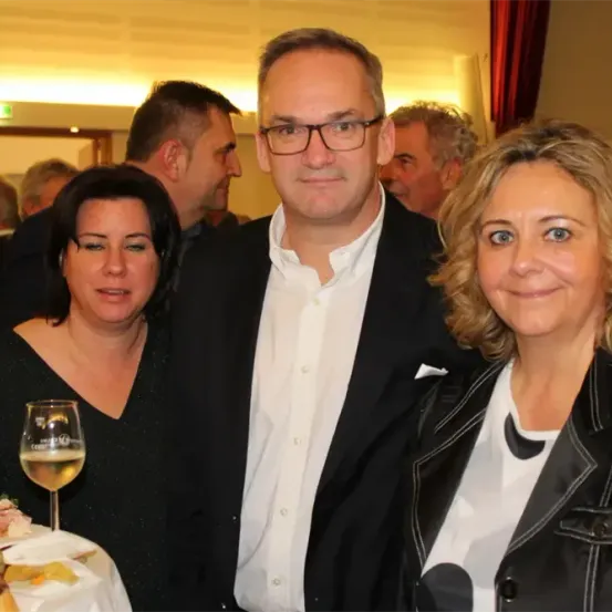 Drei Menschen stehen bei einer Veranstaltung eng beieinander. Ein Mann mit Brille, eine lächelnde Frau und eine weitere Frau, alle schauen nach vorne. Dahinter sind mehrere Menschen anwesend. Ein Tisch mit Weingläsern und Essen steht davor.