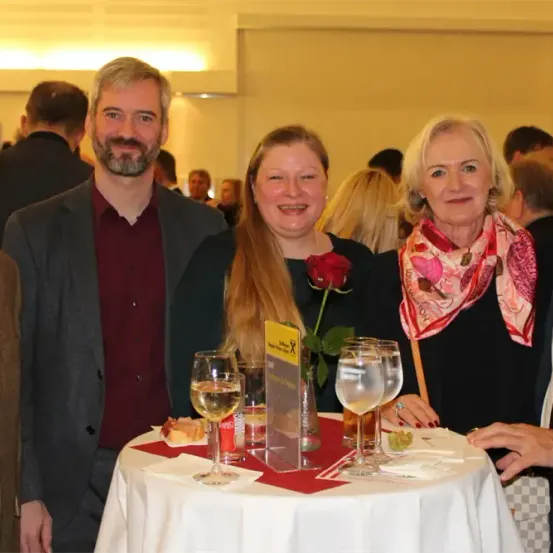 Fünf Personen stehen an einem Tisch mit Getränken. Eine Frau hält eine Rose, und eine andere Frau trägt ein Tuch. Ein Mann hält eine Handtasche.