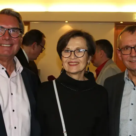 Drei Menschen lächeln und posieren für ein Foto. Die Frau in der Mitte trägt eine Brille und ein schwarzes Oberteil. Hinter ihr stehen zwei Männer. Einer trägt eine Brille und ein weißes Hemd, der andere ein kariertes Hemd.