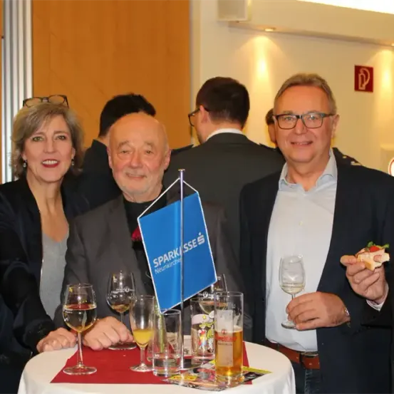 Eine Gruppe von Menschen, einschließlich eines älteren Mannes, steht um einen Tisch mit Gläsern und Essen. Ein Mann hält ein Glas Wein und ein Sandwich. Hinter ihnen steht ein weiterer Mann mit Brille und einer blauen Flagge.