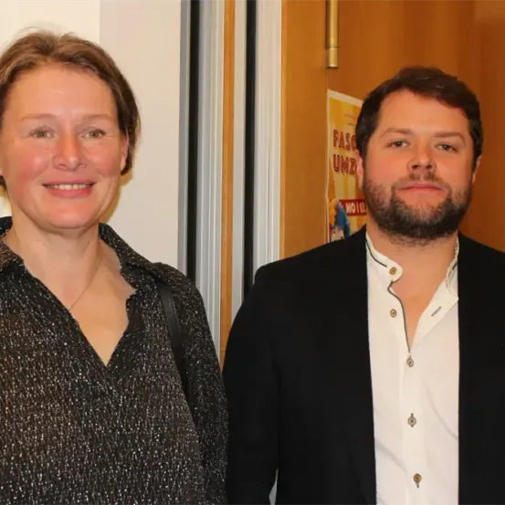 Zwei Personen stehen nah beieinander und lächeln. Eine Frau trägt ein schwarzes Oberteil mit einer Halskette. Ein Mann trägt ein weißes Hemd unter einem schwarzen Jackett. Hinter ihnen ist ein Plakat mit Text und Bild an einer Holztür angebracht.