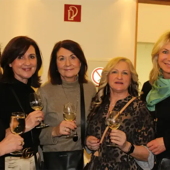 Fünf Frauen stehen in einer Indoor-Umgebung, lächeln und halten Weingläser. Eine Frau trägt ein grünes Tuch. Hinter ihnen hat eine Wand ein Feuerlöscher-Schild und ein Rauchverbotsschild.