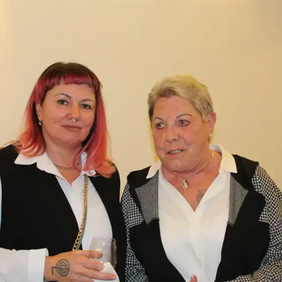 Zwei Frauen stehen nebeneinander und lächeln. Die Frau auf der linken Seite hält ein Weinglas. Die Frau auf der rechten Seite hat eine Halskette.