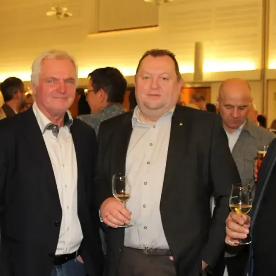 Mehrere Männer in Anzügen posieren für ein Foto, während sie Weingläser halten. Sie stehen in einem Raum mit mehreren Personen im Hintergrund.