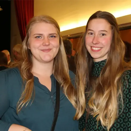 Zwei Frauen mit langen blonden Haaren lächeln und stehen zusammen. Sie befinden sich in einer Indoor-Veranstaltung. Eine trägt ein blaues Shirt, die andere ein grün gemustertes Top. Im Hintergrund sind Menschen zu sehen.