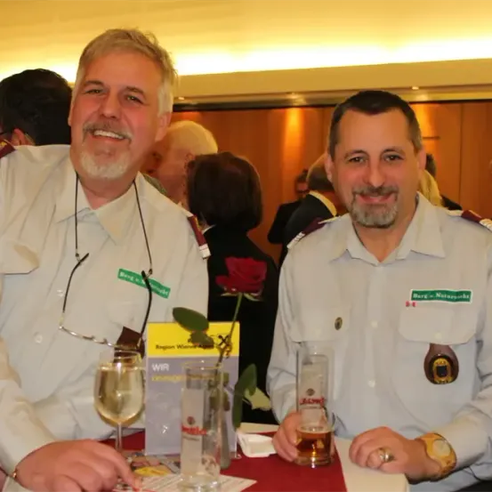 Zwei Männer in Militäruniformen lächeln in die Kamera. Sie sitzen an einem Tisch mit Gläsern Wein und Bier. Eine Rose ist zwischen ihnen platziert. Andere Leute stehen hinter ihnen.