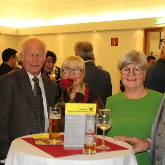 Eine Gruppe von Menschen, darunter zwei Frauen, steht um einen Tisch mit Getränken. Sie alle lächeln. Dahinter sind andere Menschen, die auch lächeln und zu sprechen scheinen.