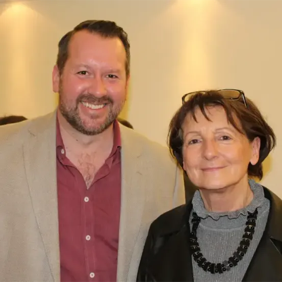 Ein Mann und eine Frau stehen lächelnd nebeneinander. Der Mann trägt ein rotes Hemd und eine helle Jacke, während die Frau eine schwarze Jacke, ein graues Rollkragenshirt und eine schwarze Halskette trägt. Sie scheinen bei einer Veranstaltung zu sein.