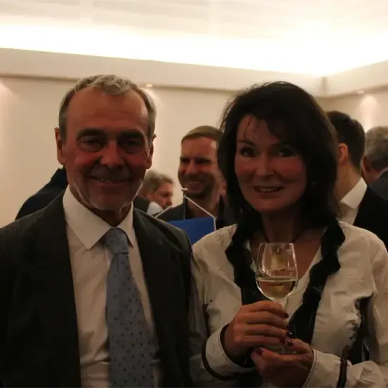 Ein Mann und eine Frau lächeln und posieren für ein Foto bei einer Veranstaltung. Der Mann trägt einen Anzug und eine Krawatte. Die Frau hält ein Glas Wein in der Hand. Dahinter stehen einige Leute und lächeln. Der Raum ist gut beleuchtet.