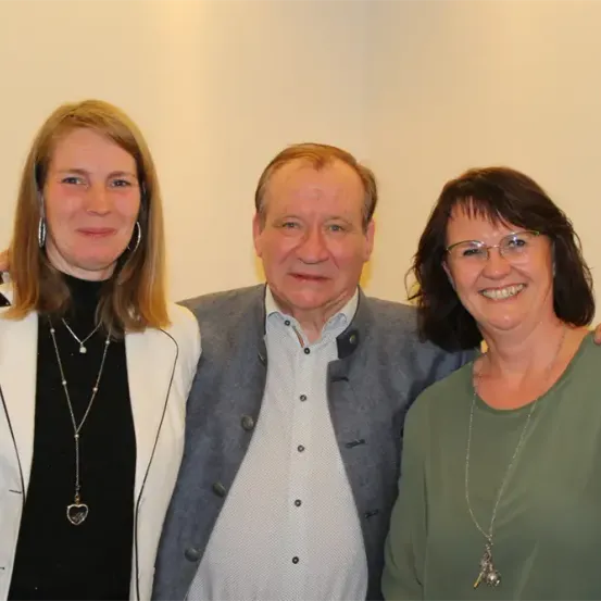Drei Personen posieren für ein Foto vor weißem Hintergrund. Die Frau auf der linken Seite trägt eine weiße Jacke, eine Halskette und Ohrringe. Der Mann in der Mitte trägt einen grauen Blazer und ein weißes Hemd. Die Frau auf der rechten Seite trägt eine Brille, ein grünes Oberteil und eine Halskette.