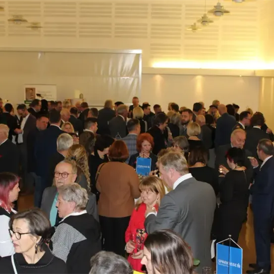 Eine große Menschenmenge ist drinnen und versammelt sich in einem Raum. Sie sind formell gekleidet, wahrscheinlich für eine Veranstaltung. Einige halten Gläser, andere unterhalten sich. Ein blaues Schild ist auf der rechten Seite sichtbar.