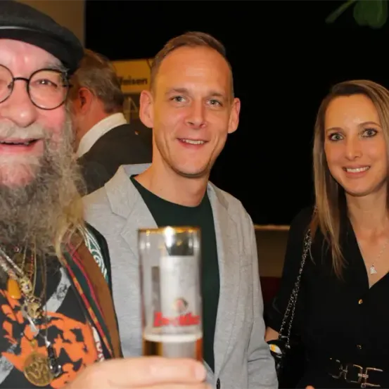 Ein Mann mit Bart und Hut hält ein Bierglas mit der Marke Budweiser. Neben ihm lächelt ein Mann in einem grauen Jackett. Hinter ihnen schaut eine Frau mit blonden Haaren zur Kamera.