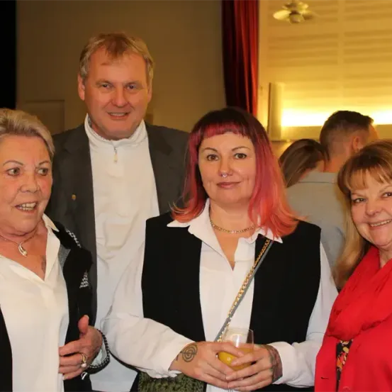 Vier Personen posieren für ein Foto bei einer Veranstaltung; die beiden Frauen an den Seiten lächeln, während die zentrale Frau mit rosa Haaren ein Getränk in der Hand hält. Ein Mann steht hinter ihnen und lächelt.