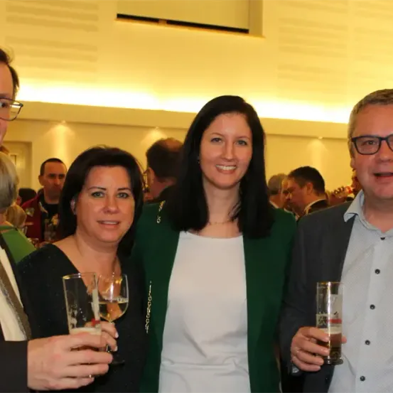 Vier Menschen lächeln und posieren für ein Foto, während sie Getränke in der Hand halten, in einem Raum mit mehreren Menschen im Hintergrund.