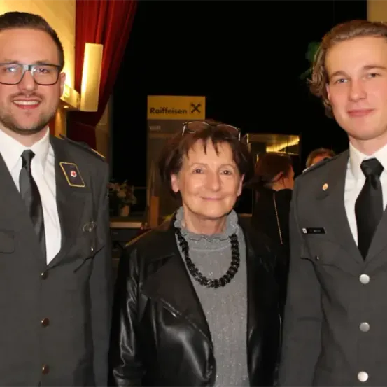 Drei Personen stehen eng beieinander und lächeln für ein Foto. Der Mann links trägt eine Militäruniform mit einem roten Kreuz auf der Schulter. Der Mann rechts trägt eine ähnliche Uniform. Die Frau in der Mitte trägt eine schwarze Lederjacke.
