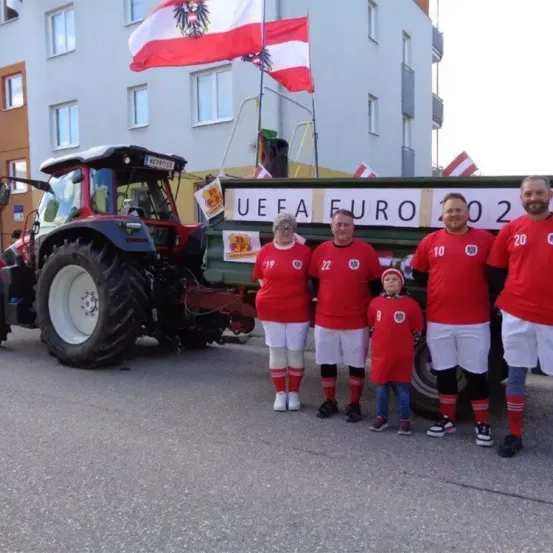 Fünf Personen, darunter ein Kind, in roten Trikots stehen vor einem Traktor mit dem Schriftzug UEFA EURO 2020. Dahinter ist ein Gebäude mit mehreren Fenstern und Flaggen zu sehen.