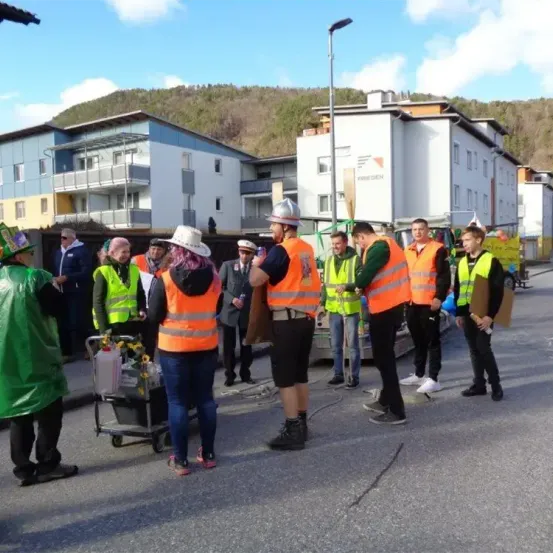 Eine Gruppe von Menschen in Sicherheitswesten steht auf der Straße und unterhält sich. Ein Mensch mit Helm und ein Mann mit einem Trolley sind ebenfalls anwesend. Ein weißes Gebäude mit der Aufschrift 'FRIEDEN' befindet sich hinter ihnen.