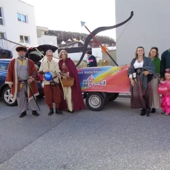 Eine Gruppe von Menschen in mittelalterlichen Kostümen posiert für ein Foto. Sie stehen vor einem dekorierten Anhänger mit einem Banner. Im Hintergrund sind Gebäude und ein Berg zu sehen.