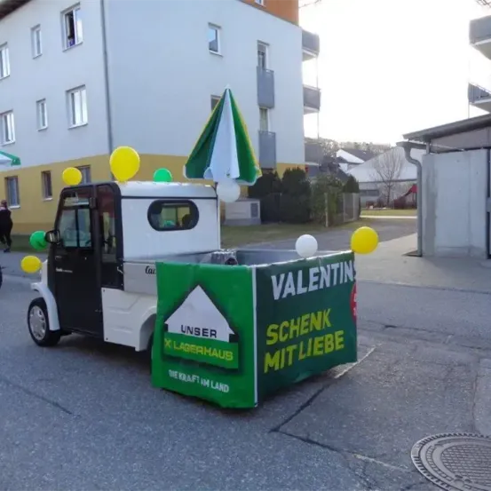 Ein dekoriertes Elektrofahrzeug zeigt ein Schild mit der Aufschrift 'Valentin Schenk mit Liebe' und hat Luftballons und eine Fahne darauf, geparkt in einer Straße.