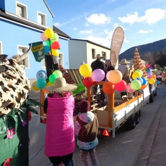Ein bunter Umzug auf der Straße mit einem geschmückten Wagen voller Luftballons und Menschen in Kostümen. Dahinter ein Gebäude mit einem Schild, das 'Jackpot' steht.