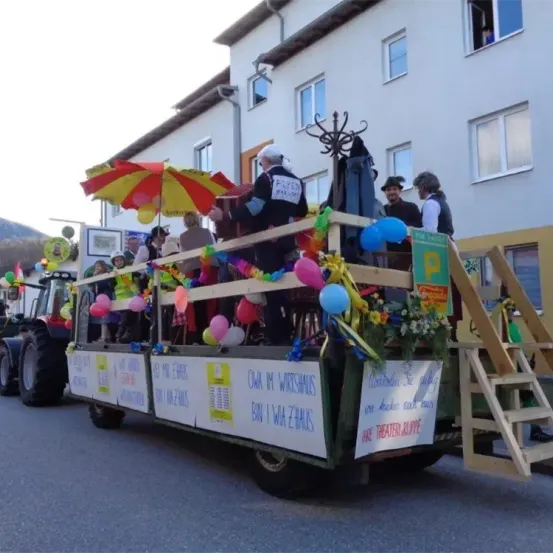 Ein geschmückter Paradewagen mit Dekorationen, Luftballons und Menschen, gezogen von einem Traktor, vor einem Gebäude mit mehreren Fenstern.