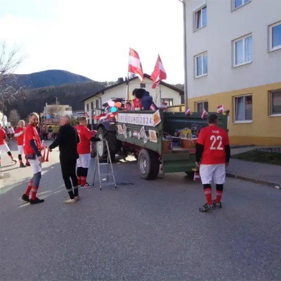 Mehrere Menschen in roten Trikots und Shorts versammeln sich um einen Lastwagen mit Flaggen und Schildern für die UEFA EURO 2024. Eine Person steht im Lastwagen, und ein Gebäude mit Glasfenstern befindet sich dahinter.