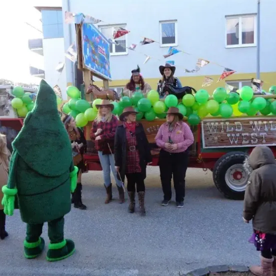 Ein Festwagen mit Menschen in Cowboy-Hüten und grünen Luftballons. Vorne steht eine Person in einem grünen Kostüm.