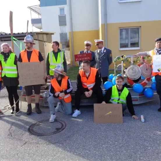 Eine Gruppe von Menschen in orangefarbenen Warnwesten posiert für ein Foto vor einem Gebäude. Einige halten Schilder und Helme. Dahinter hält ein Mann ein Schild mit der Aufschrift 'Wille Flaschen.' Die Umgebung umfasst Luftballons und einen Feuerlöscher.