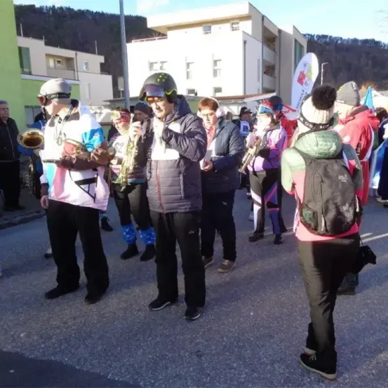 Eine Gruppe von Menschen in Kostümen und Helmen steht auf einer Straße. Sie halten Musikinstrumente in den Händen. Sie marschieren wahrscheinlich bei einer Parade mit. Die Gebäude sind hinter ihnen.