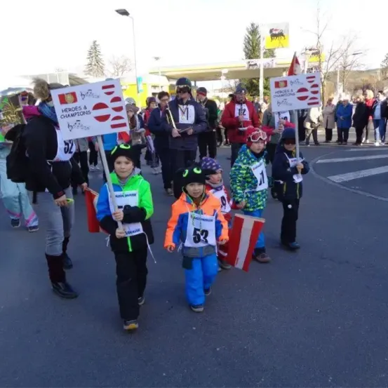 Mehrere Kinder, jedes mit einer Nummer auf dem Shirt, laufen in einer Parade. Sie halten Schilder mit der Aufschrift Helden und Champions. Eine Menschenmenge steht an der Seite.