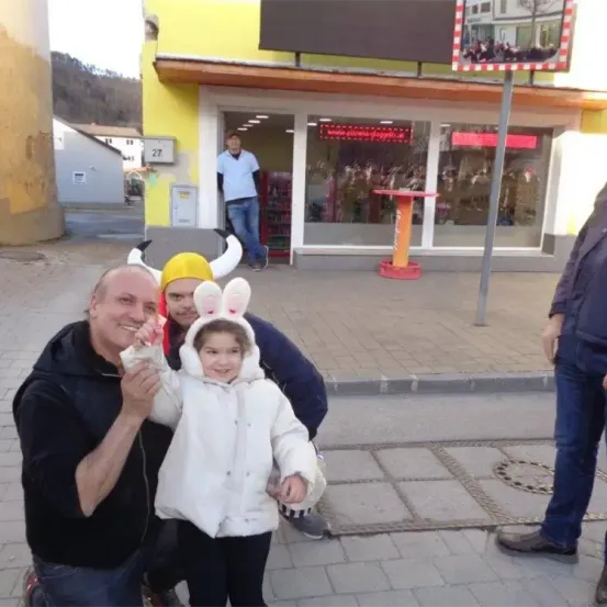 Drei Personen posieren für ein Foto auf der Straße, mit einem Gebäude und einem Mann im Hintergrund. Der Mann trägt einen Wikingerhut und die anderen sind in Kostümen gekleidet.